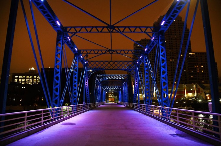 A pedestrian's view from Blue Bridge at night with the buildings of downtown Grand Rapids lit up in the background