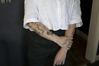 Vanessa DeCouto, wearing a white button-up shirt with the sleeves rolled up, shows off tattoos on their forearms and hands.